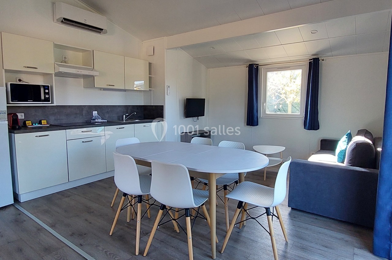 Cuisine ouverte avec table ronde, chaises blanches, canapé gris, réfrigérateur, micro-ondes et fenêtre lumineuse.