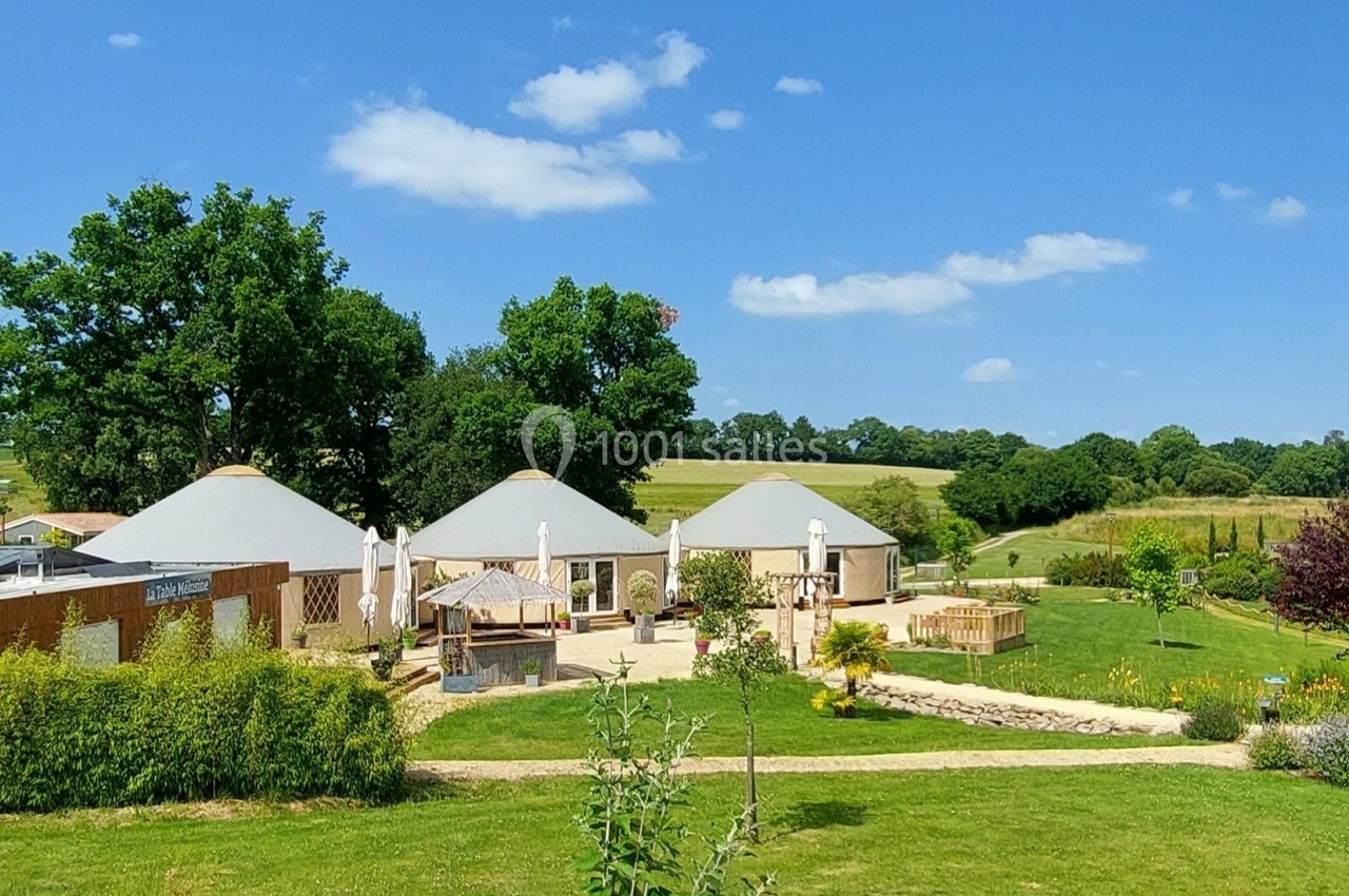 Vue d'un espace extérieur avec des bâtiments circulaires, terrasse, parasols et jardin verdoyant sous un ciel bleu.