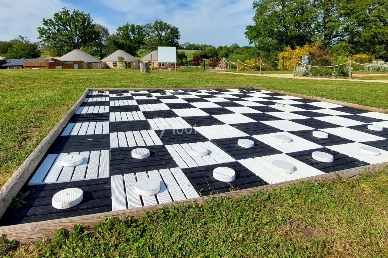 Jeu de dames géant en bois noir et blanc installé sur une pelouse, entouré d'arbres et de bâtiments.