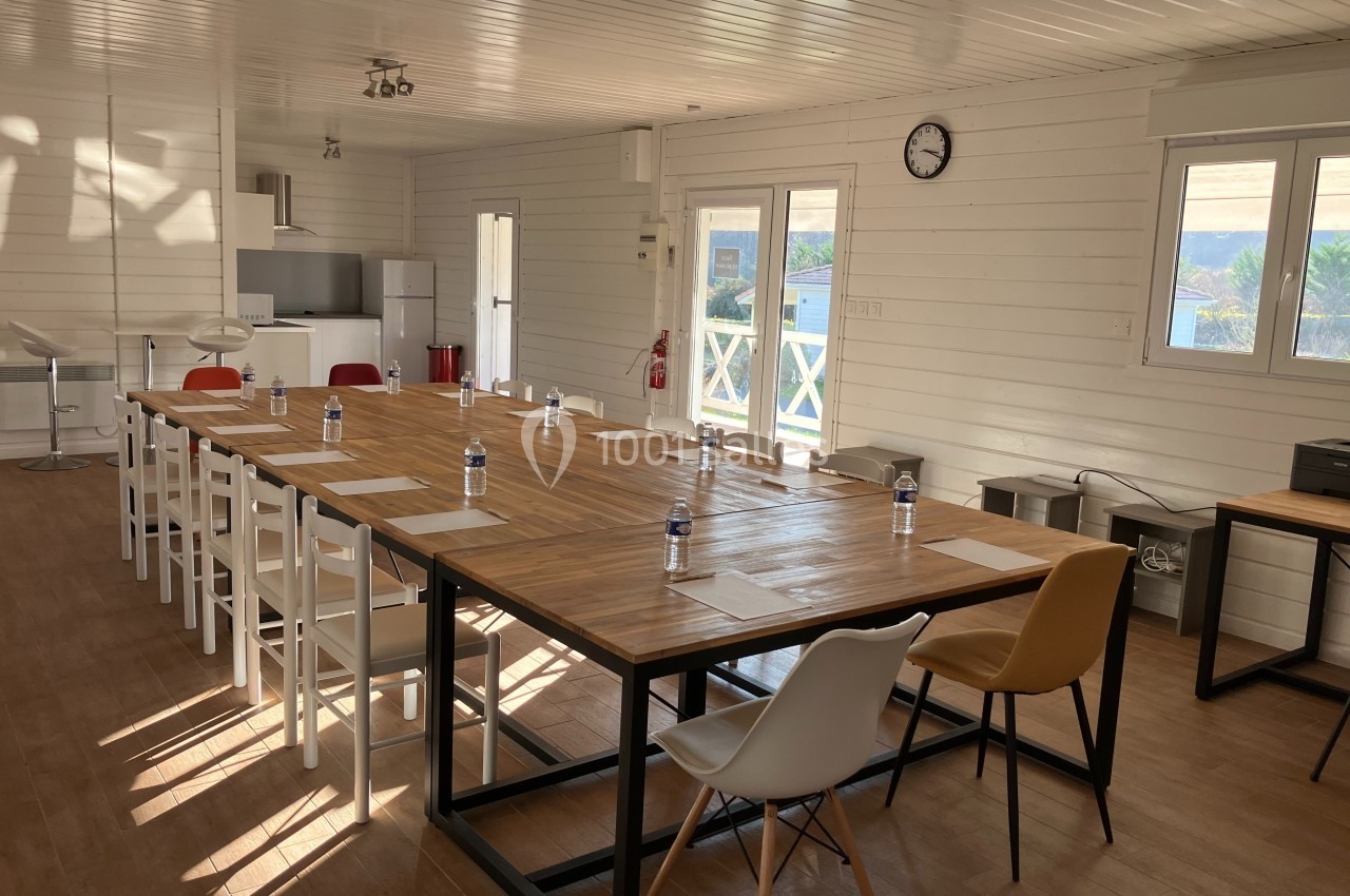 Salle lumineuse avec de grandes tables en bois, des chaises variées, des bouteilles d'eau et des feuilles disposées.