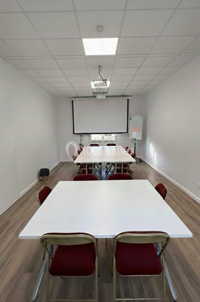 Salle de réunion lumineuse avec tables blanches, chaises rouges, projecteur, écran et tableau blanc.