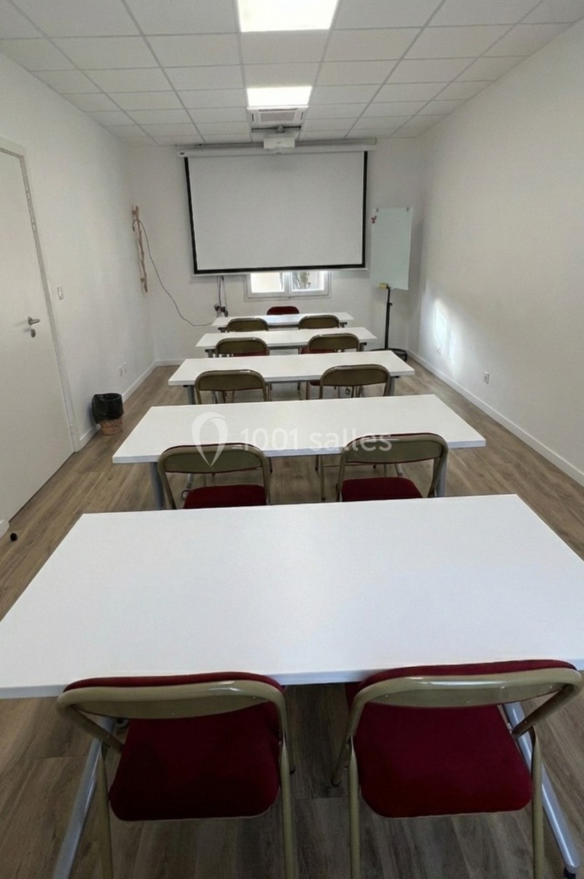 Salle de formation avec tables blanches alignées, chaises rouges, tableau blanc et écran de projection.