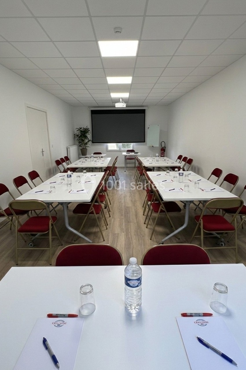 Salle de réunion lumineuse avec tables en U, chaises rouges, écran de projection et fournitures de bureau.