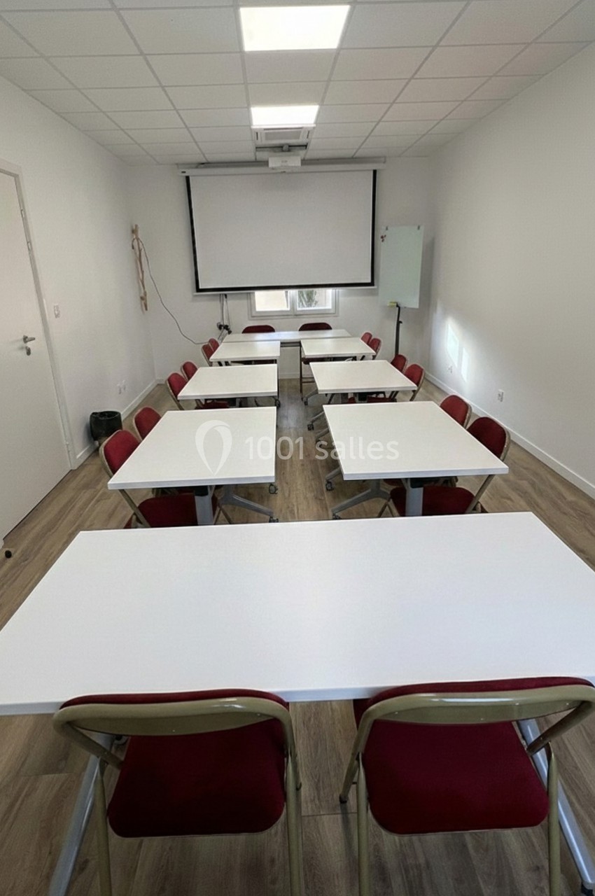 Salle de réunion lumineuse avec tables blanches, chaises rouges, tableau blanc et écran de projection.