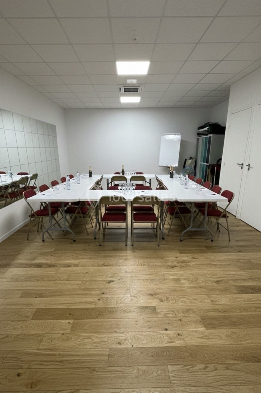 Salle de réunion avec tables disposées en rectangle, chaises rouges, tableau blanc et miroir sur un mur.