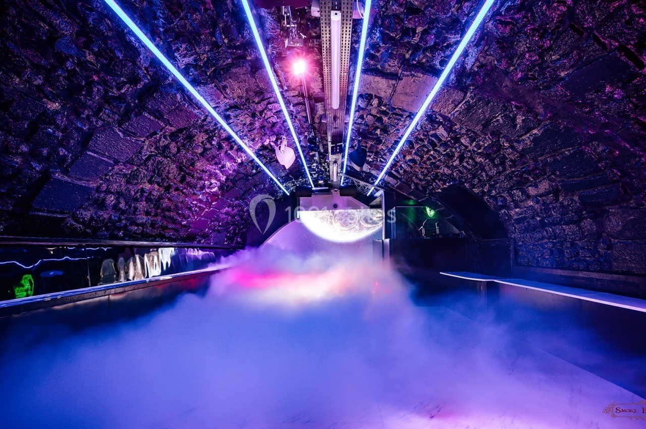 Tunnel en pierre éclairé par des lumières néon violettes et bleues, avec de la fumée au sol et un projecteur central.