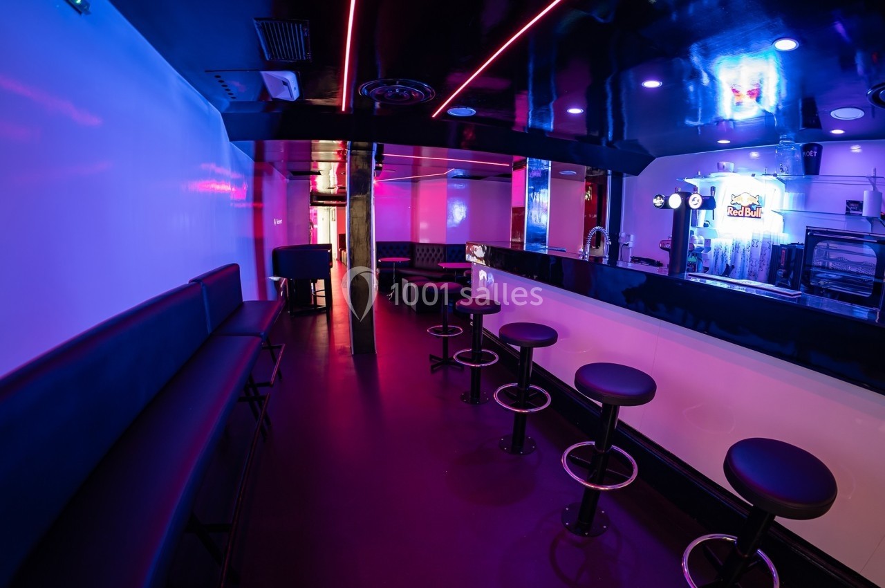 Bar moderne avec comptoir blanc, tabourets noirs, éclairage néon rose et bleu, et banquettes le long des murs.