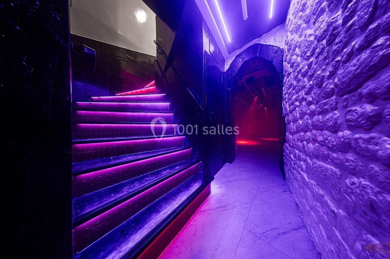 Escalier éclairé par des néons violets menant à un couloir en pierre dans une ambiance tamisée.