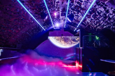 Salle voûtée en pierre avec éclairage LED coloré, mobilier minimaliste et comptoir de bar au fond.