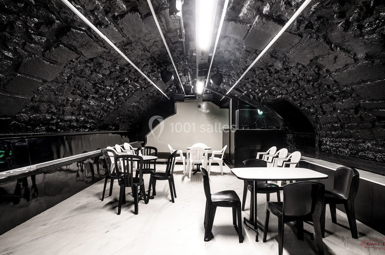 Salle voûtée avec murs en pierre sombre, éclairage néon et mobilier moderne composé de tables blanches et chaises noires.