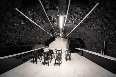 Salle voûtée en pierre avec éclairage LED coloré, mobilier minimaliste et comptoir de bar au fond.