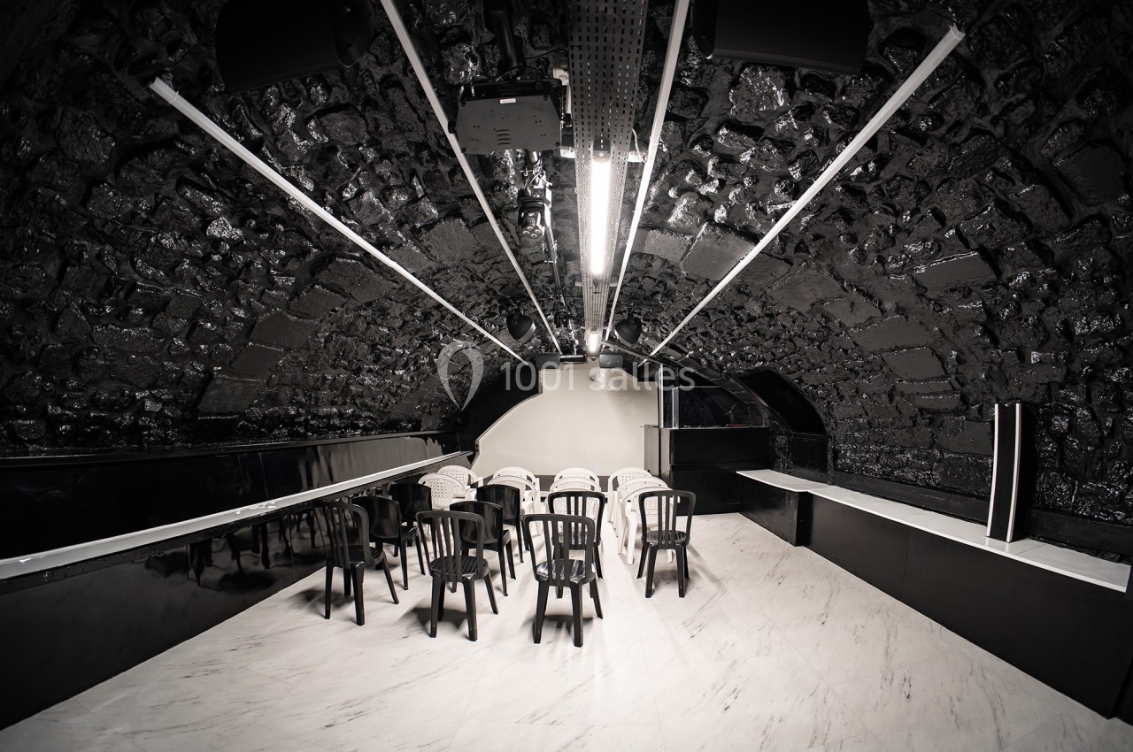 Salle voûtée en pierre sombre avec des chaises noires disposées face à un écran blanc.