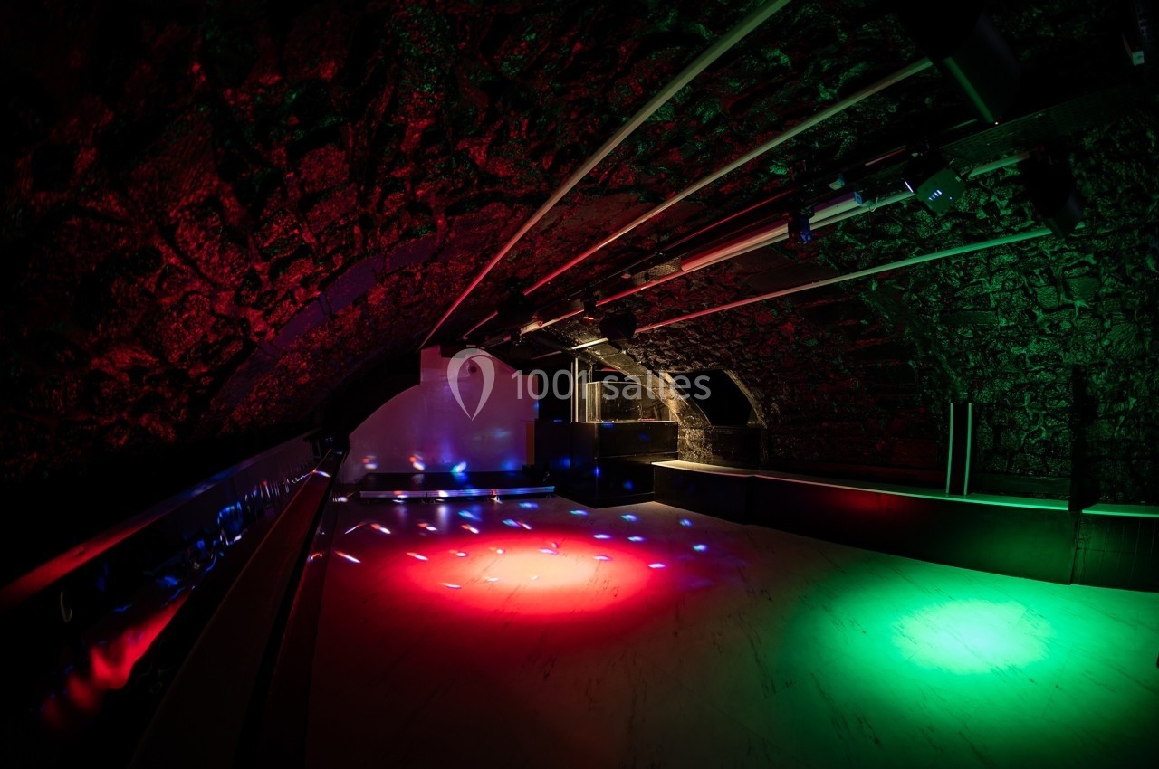Salle voûtée avec éclairage coloré rouge et vert, sol clair et murs en pierre sombre.