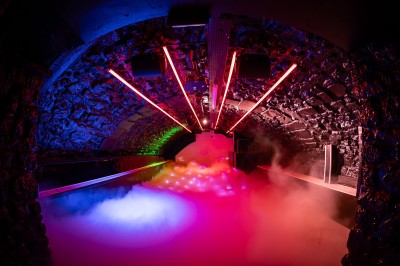 Salle voûtée en pierre avec éclairage LED coloré, mobilier minimaliste et comptoir de bar au fond.