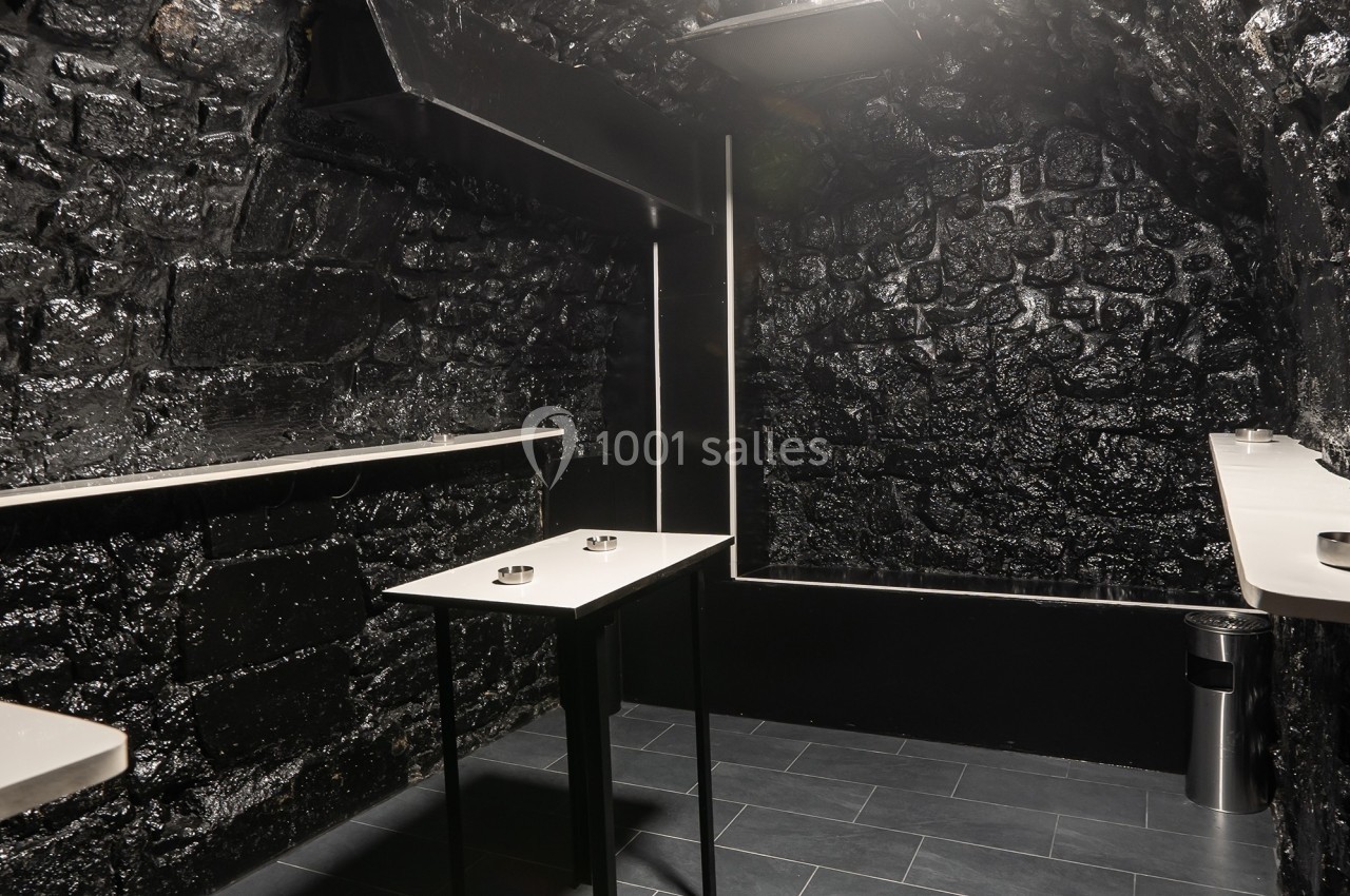 Salle sombre avec murs en pierre noire, tables hautes blanches et sol en carrelage gris.