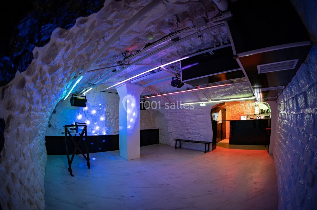 Salle voûtée en pierre blanche avec éclairage coloré, piste de danse vide et comptoir de bar au fond.