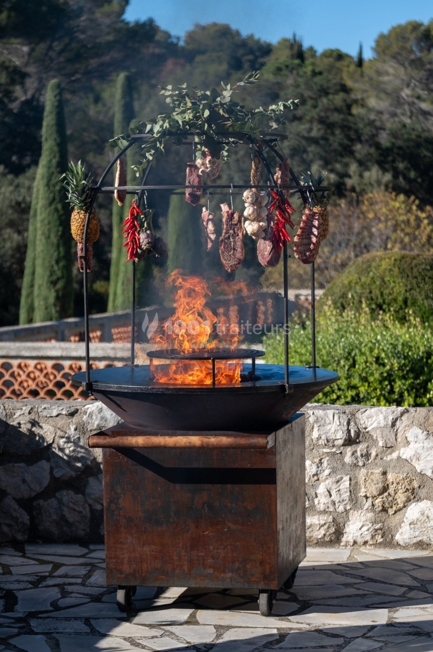 Barbecue extérieur avec feu vif, viandes, piments et ananas suspendus, entouré de végétation méditerranéenne.