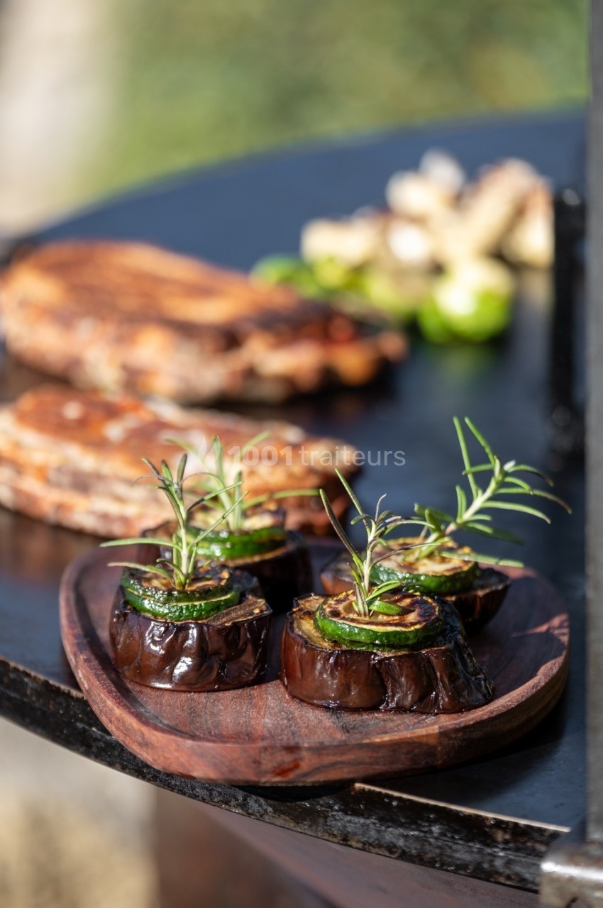Tranches d'aubergines grillées garnies de romarin sur une assiette en bois, avec des aliments cuisant en arrière-plan.