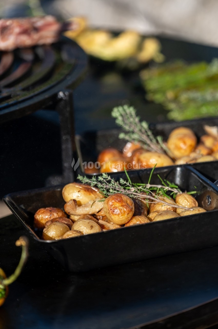 Pommes de terre rôties dans un plat noir, garnies de branches de thym, posées sur une table près d'un barbecue.