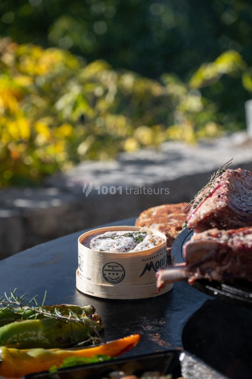 Fromage Mont d'Or fondu dans sa boîte en bois, entouré de légumes grillés et de viande, en extérieur ensoleillé.