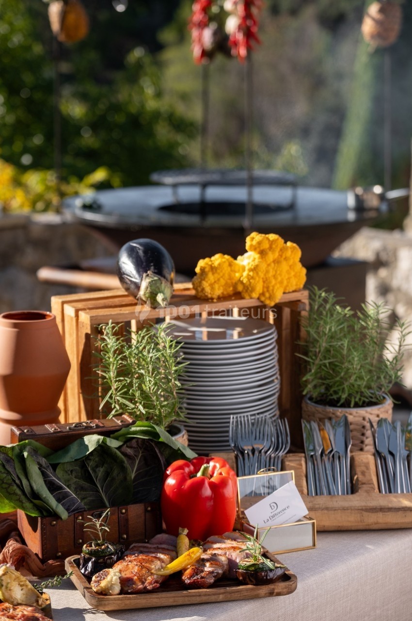 Table dressée en extérieur avec des plats cuisinés, légumes frais, herbes aromatiques et un barbecue en arrière-plan.