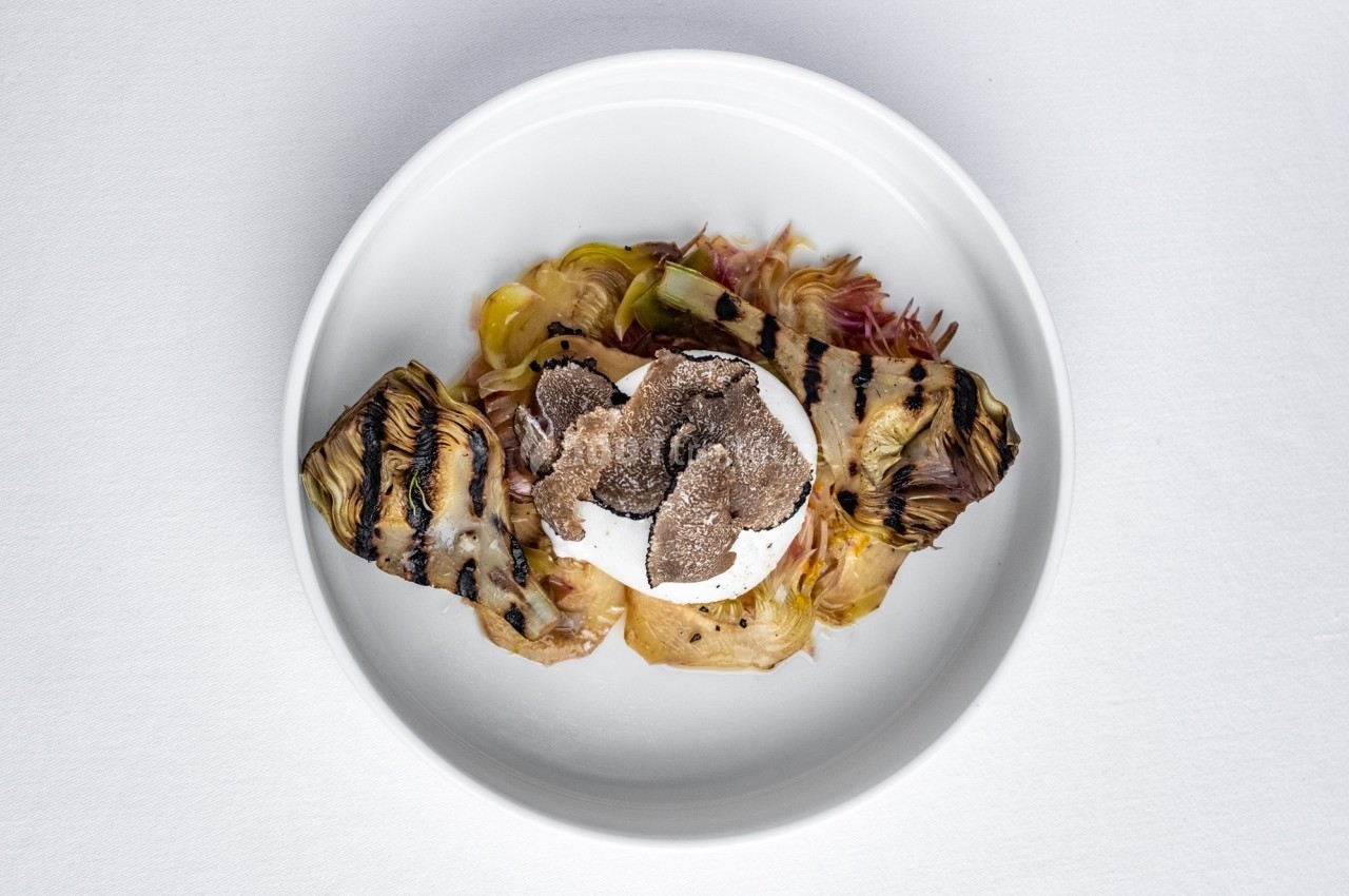 Assiette contenant des artichauts grillés, une burrata et des tranches de truffe noire, présentée sur une nappe blanche.