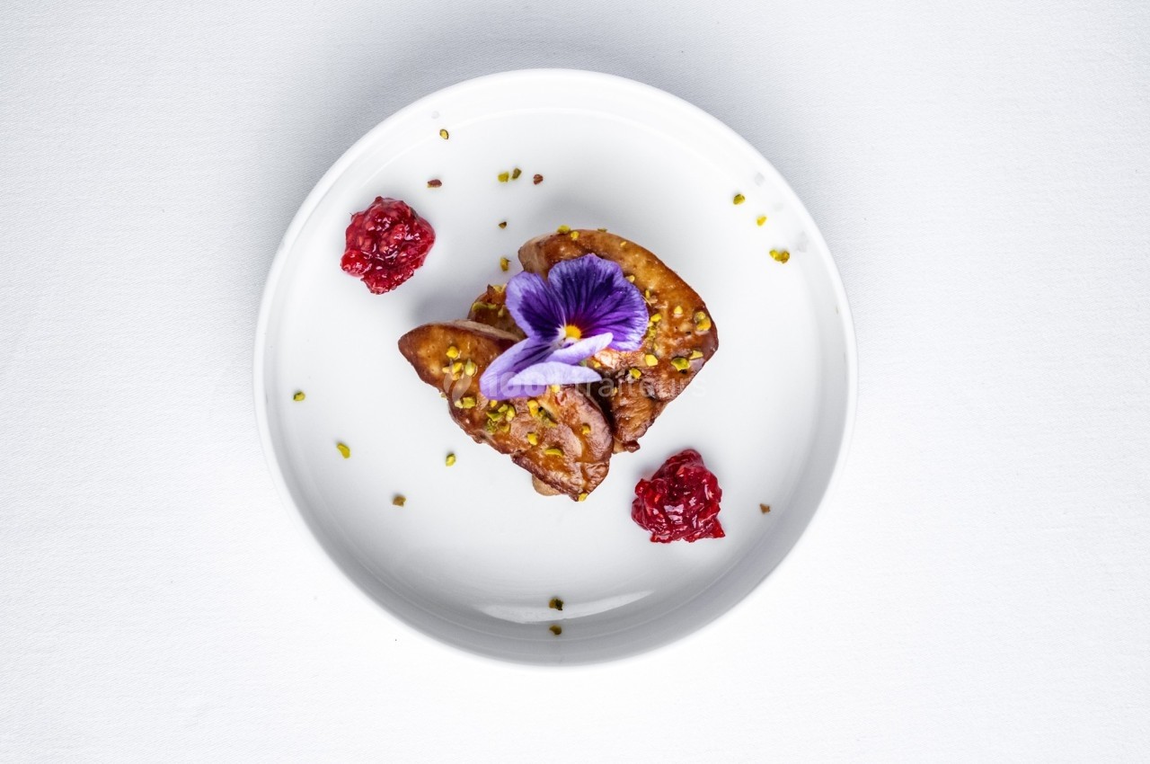 Assiette blanche contenant un plat raffiné avec foie gras, fleur comestible violette et garnitures de fruits rouges.