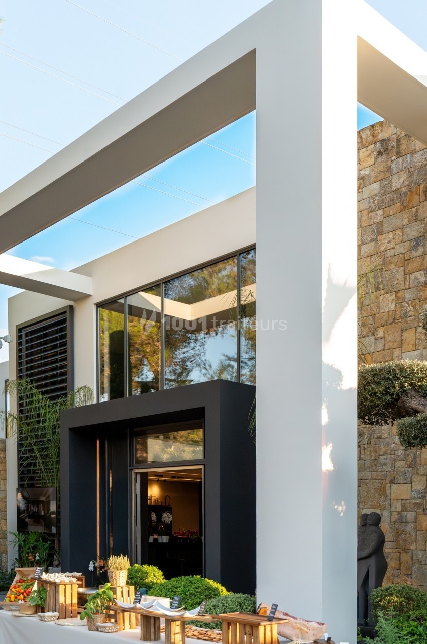 Façade moderne avec grandes baies vitrées, structure géométrique blanche et mur en pierre, entourée de végétation.