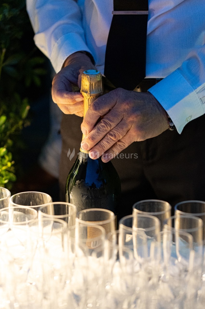 Un homme en chemise blanche ouvre une bouteille de champagne sur une table garnie de verres vides.
