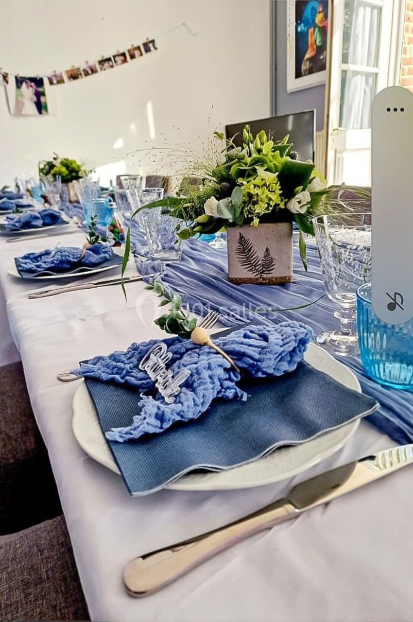 Table dressée avec une nappe blanche, serviettes bleues, vaisselle élégante et centre de table floral.