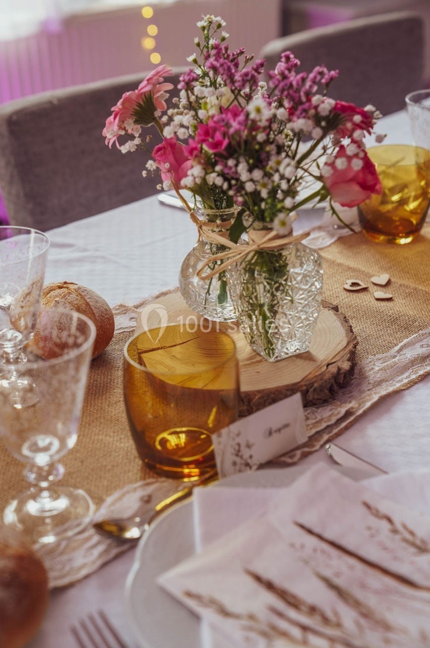 Vase en verre avec des fleurs roses et blanches sur une table décorée avec des verres ambrés et une nappe beige.