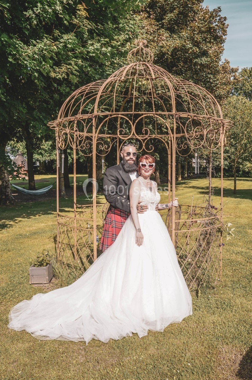 Un couple en tenue de mariage pose devant une structure en fer forgé dans un jardin verdoyant.