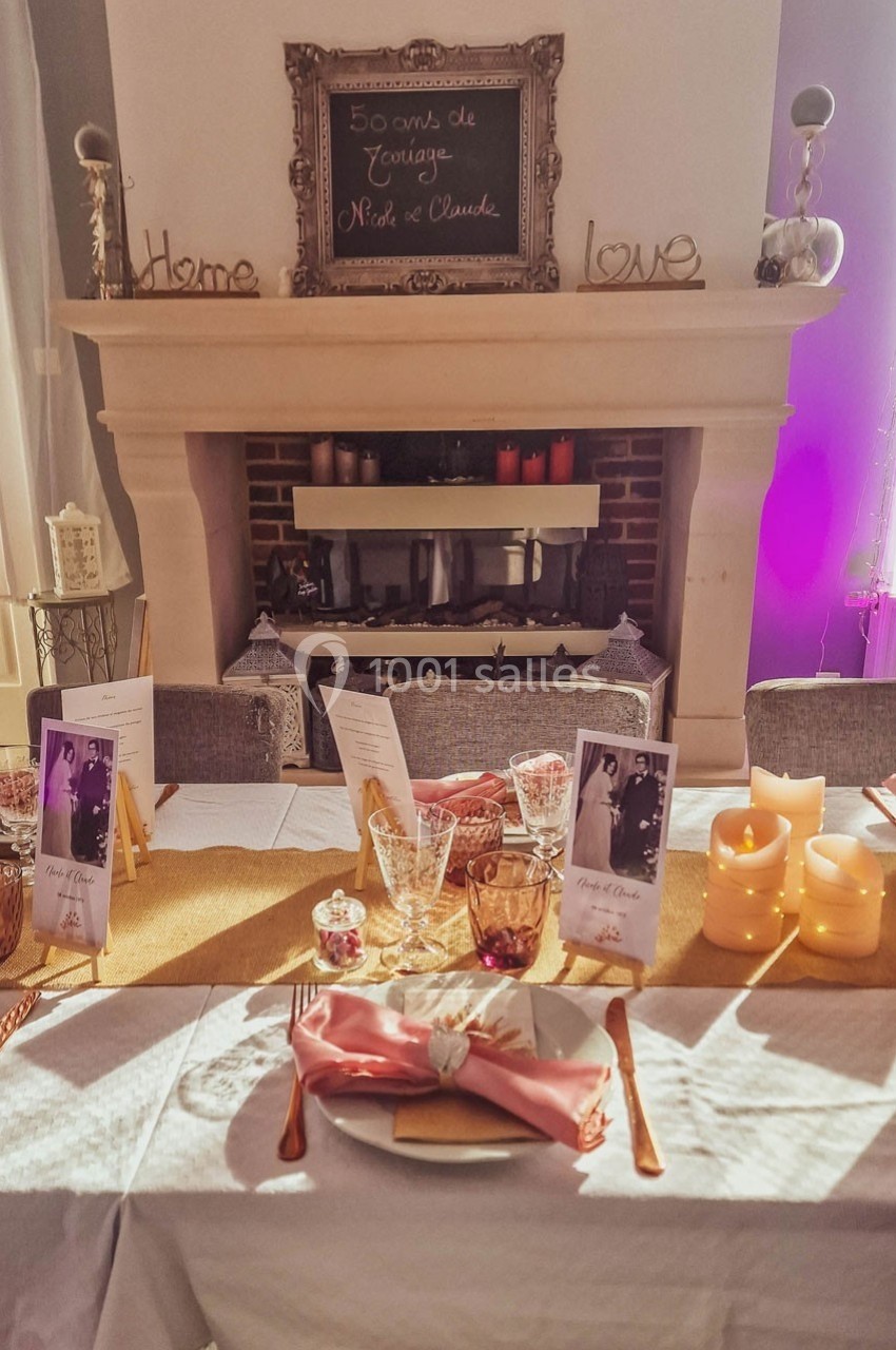 Table décorée pour une célébration, avec vaisselle élégante, bougies, photos et une cheminée en arrière-plan.