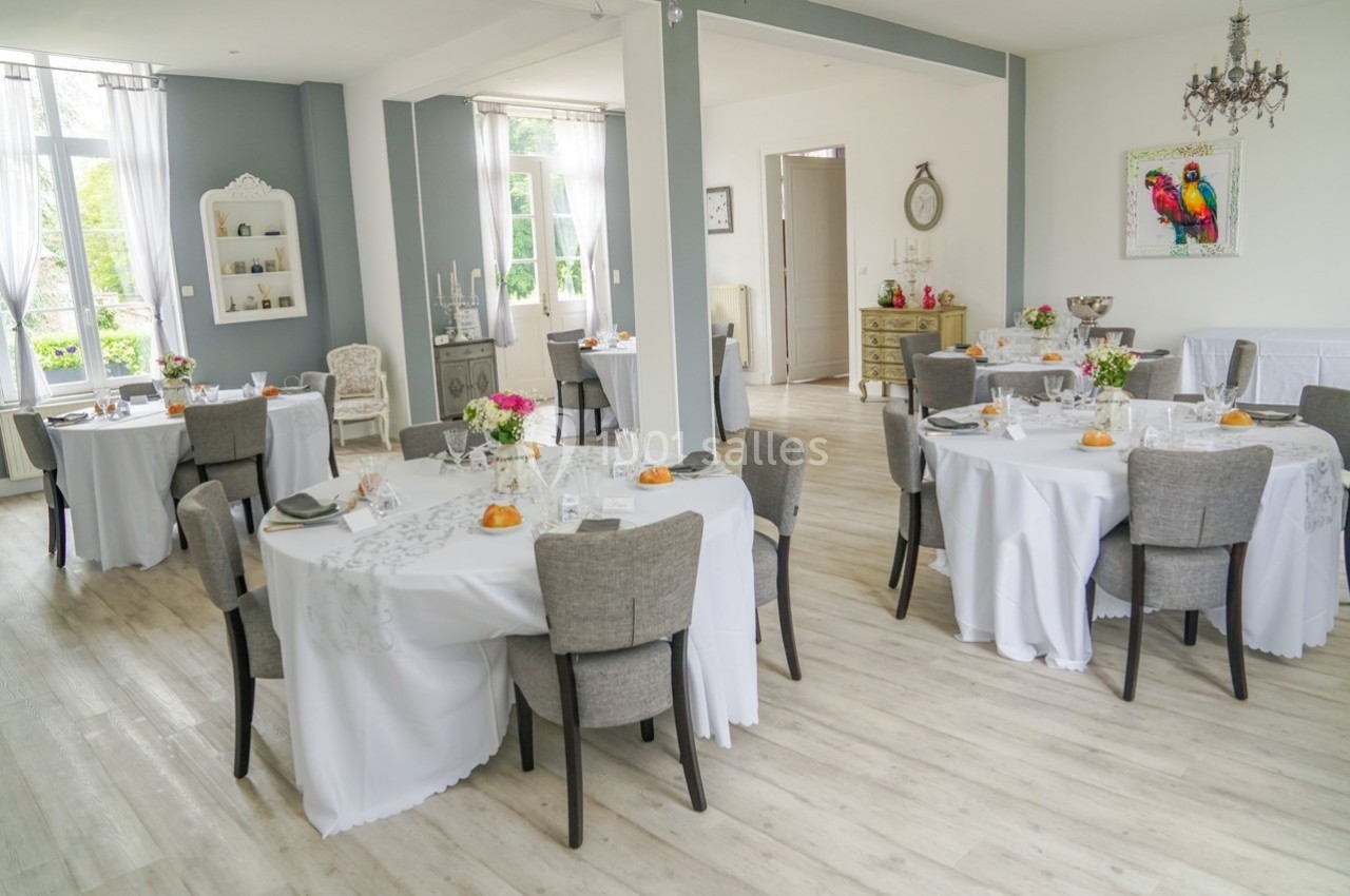 Salle lumineuse décorée pour un repas, avec tables rondes dressées, nappes blanches et arrangements floraux.