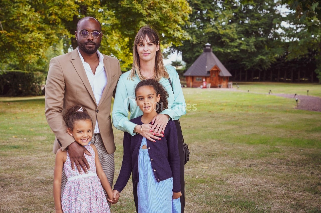 Un couple pose avec deux jeunes filles dans un parc verdoyant, avec une petite cabane en bois en arrière-plan.