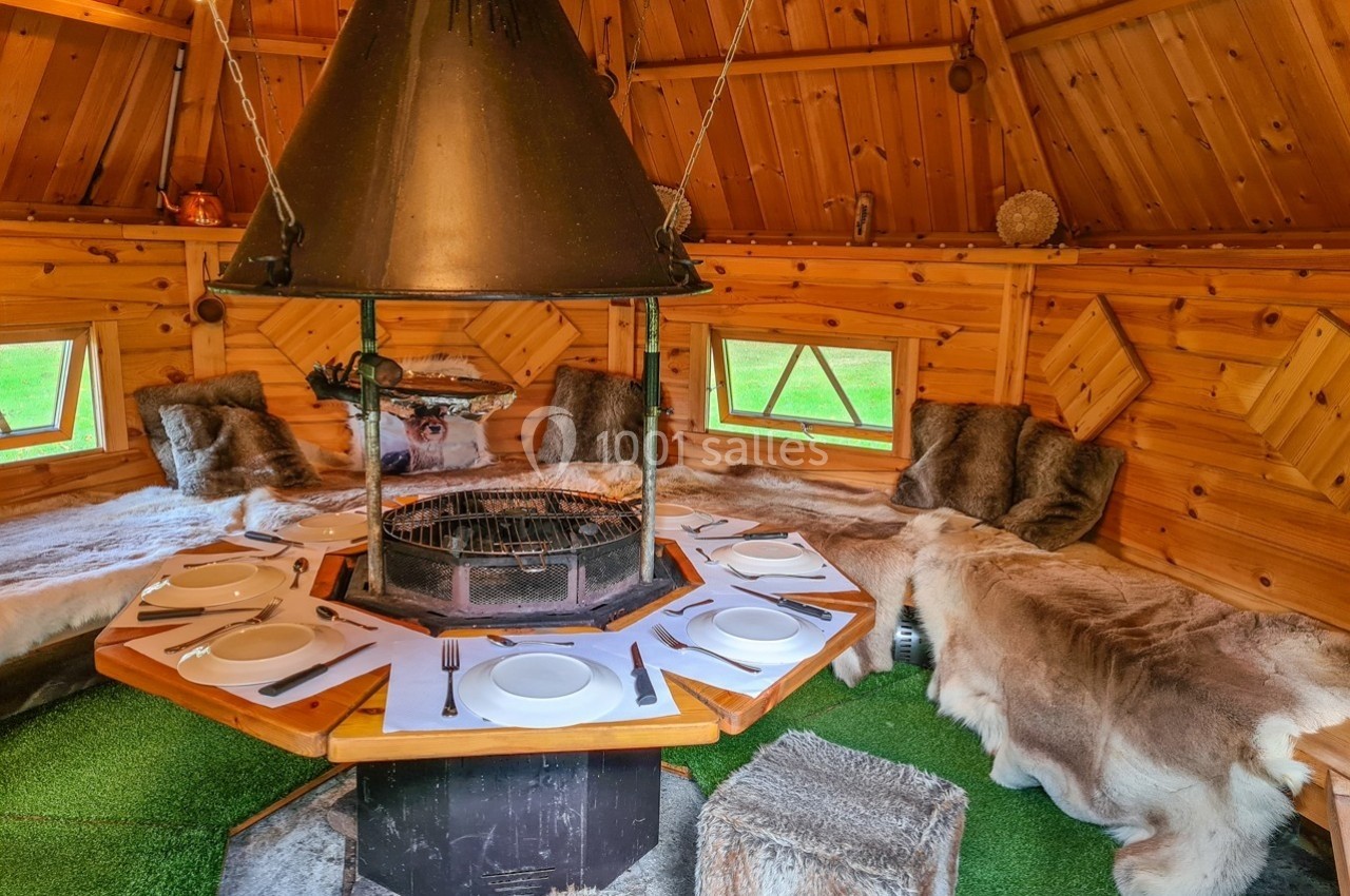 Intérieur d'un chalet en bois avec un barbecue central, des bancs recouverts de peaux et une table dressée.
