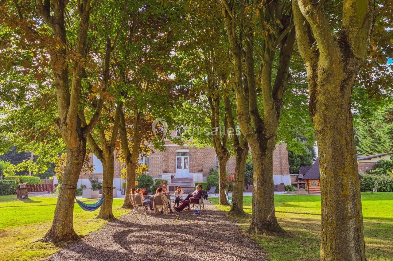 Groupe de personnes assises en cercle sous des arbres dans un jardin, avec une maison en briques en arrière-plan.
