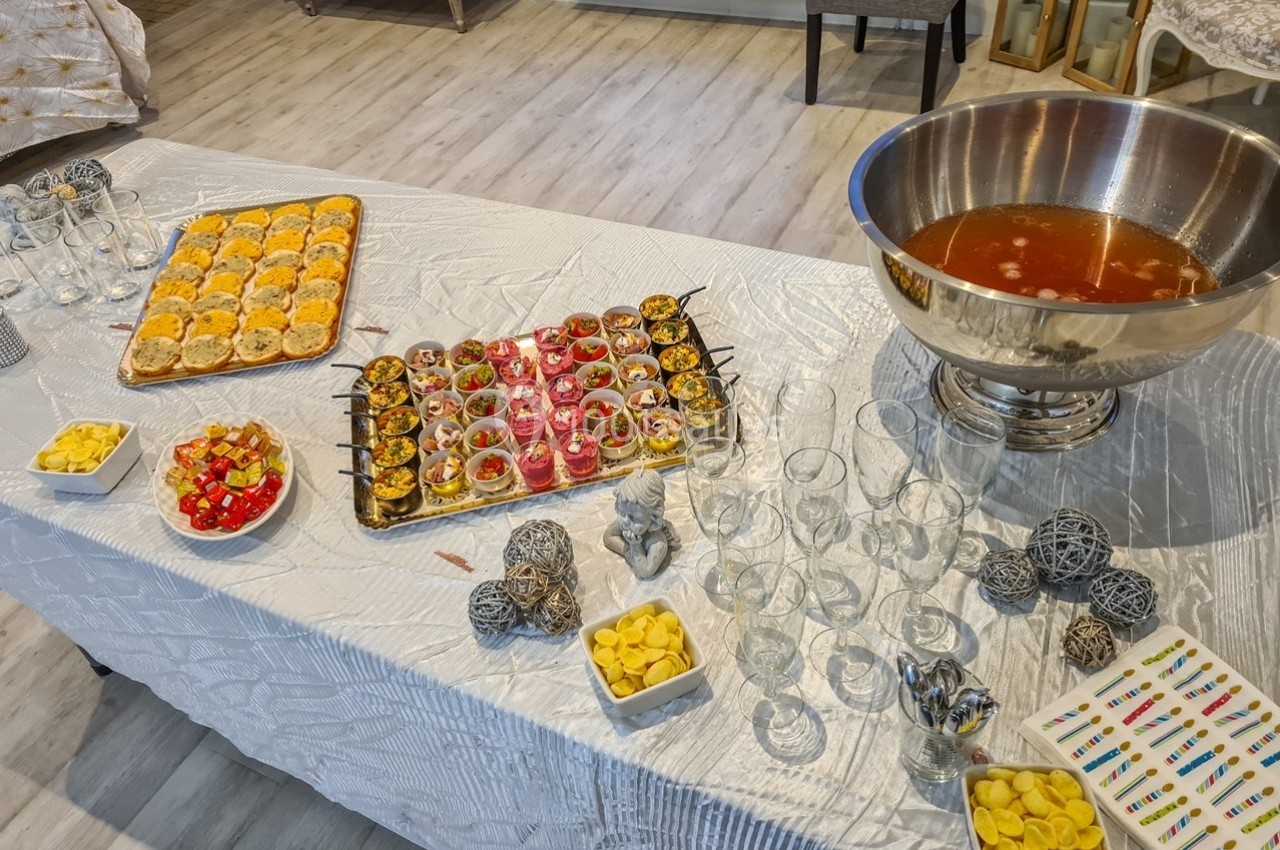 Buffet avec amuse-bouches variés, bol de punch, verres vides et décorations sur une nappe blanche.