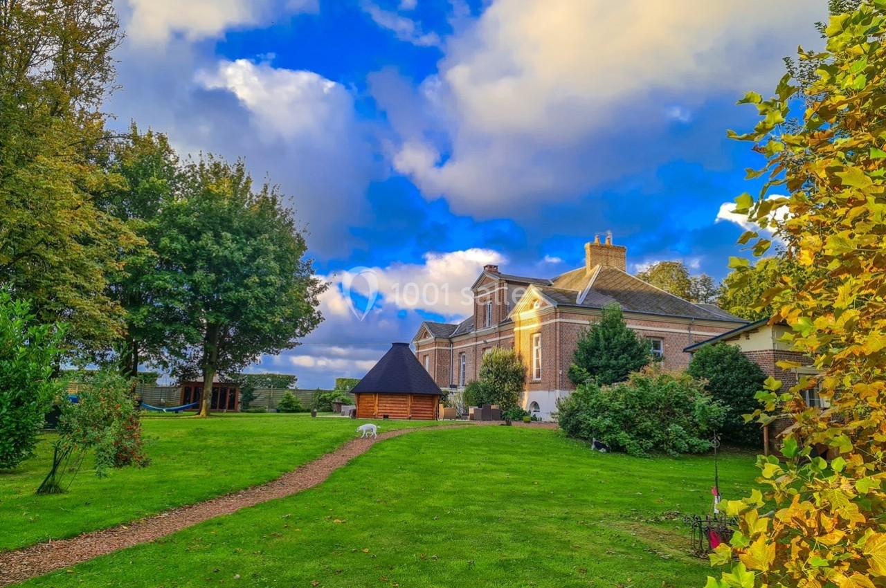 Grande maison en briques entourée d'un jardin verdoyant avec arbres, pelouse et ciel bleu partiellement nuageux.