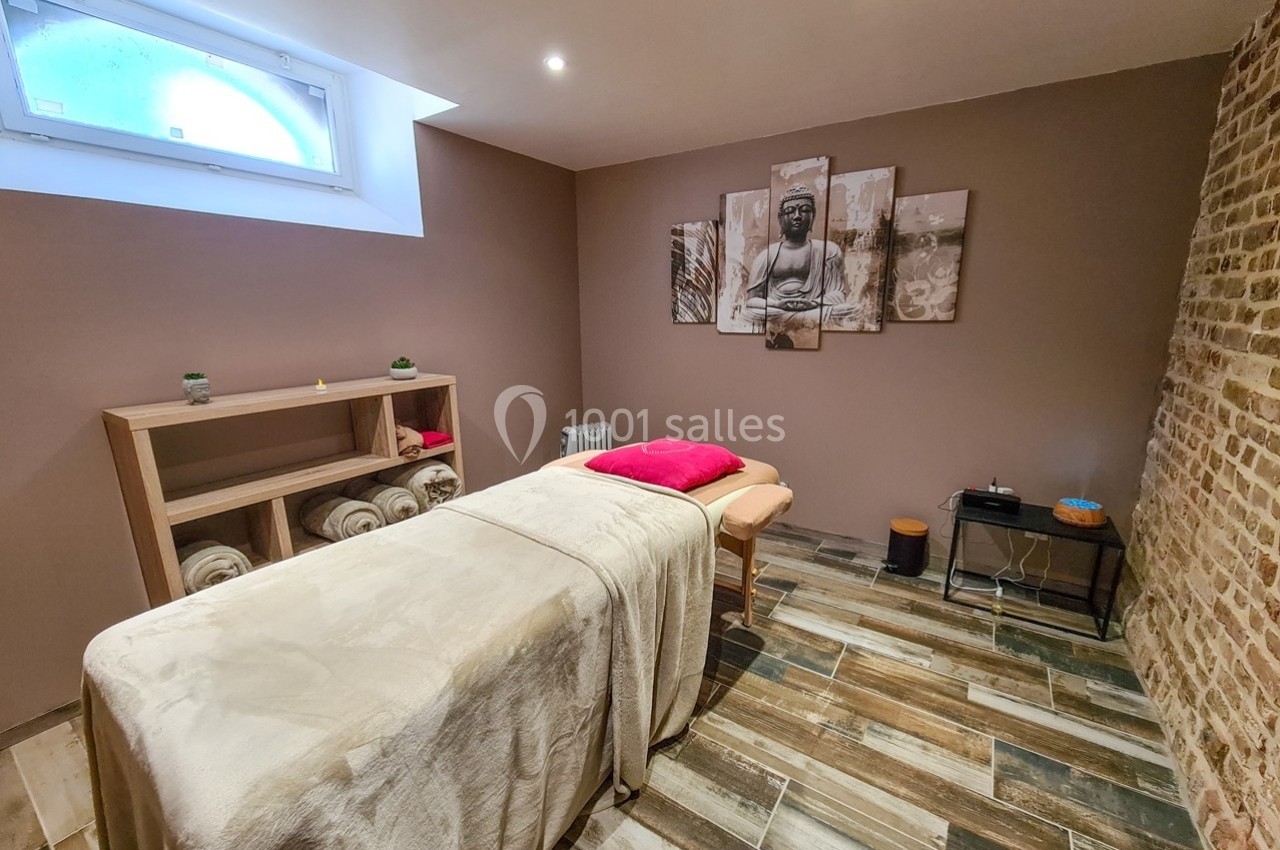 Salle de massage avec table recouverte d'un drap beige, décorée de tableaux zen et éclairée par une fenêtre en hauteur.