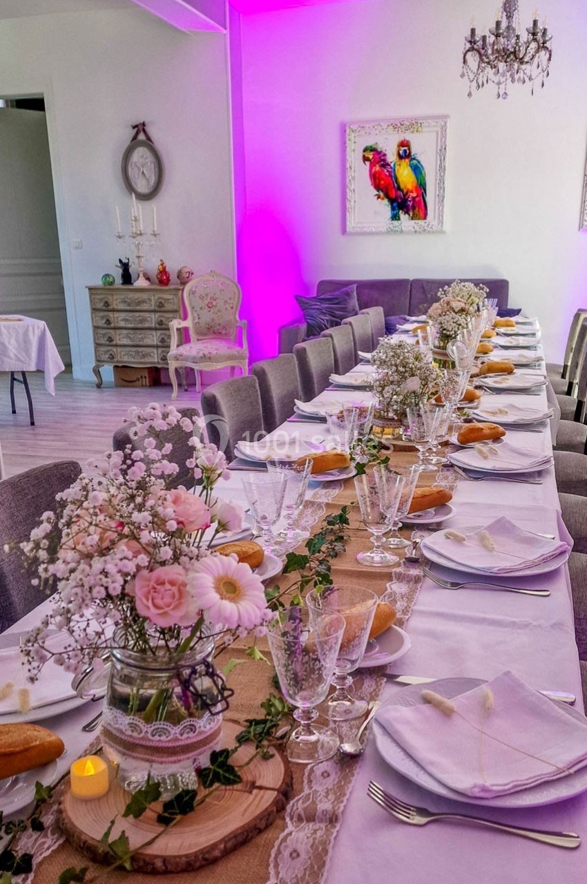 Table élégamment dressée avec nappes blanches, fleurs, bougies et vaisselle, dans une salle décorée de tons roses et violets.