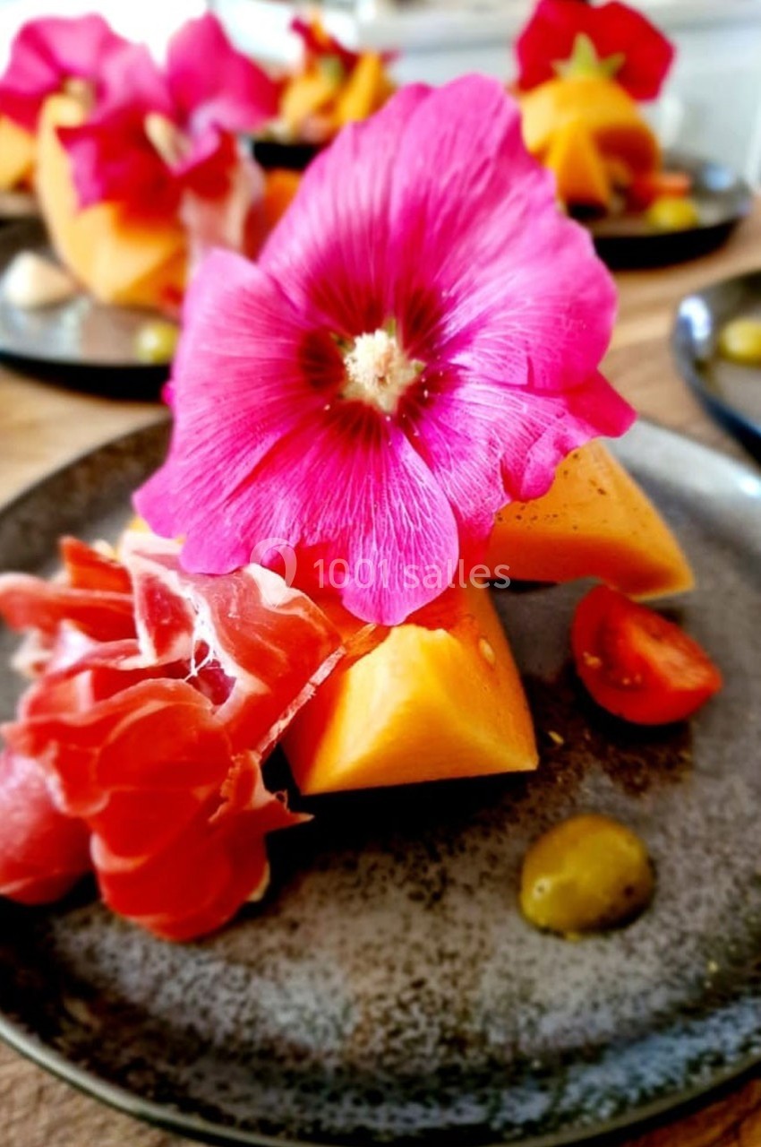 Assiette composée de tranches de melon, jambon cru, tomate cerise et fleur comestible sur une table en bois.