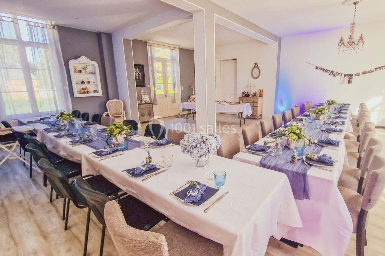 Salle lumineuse décorée pour un repas, avec deux longues tables dressées, nappes blanches et arrangements floraux.