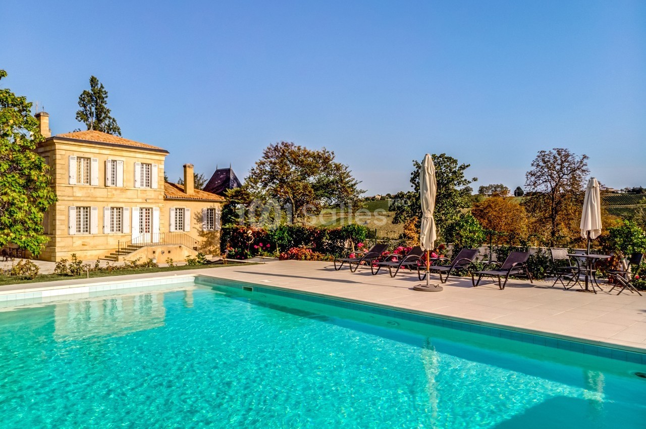 Piscine extérieure bordée de chaises longues et parasols, avec une maison en pierre et un jardin en arrière-plan.