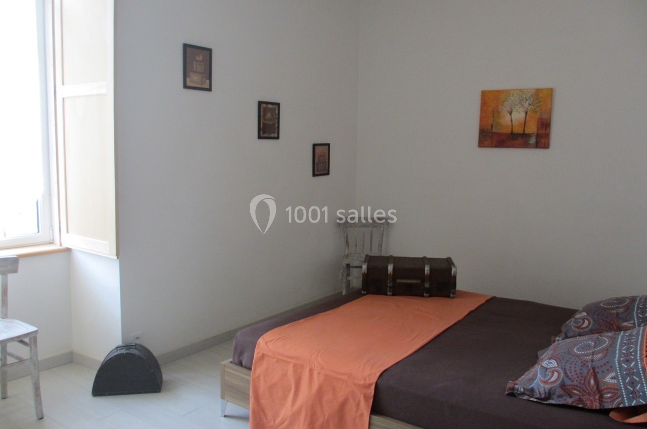 Chambre lumineuse avec lit double, drap orange, murs blancs décorés et fenêtre ouverte laissant entrer la lumière.