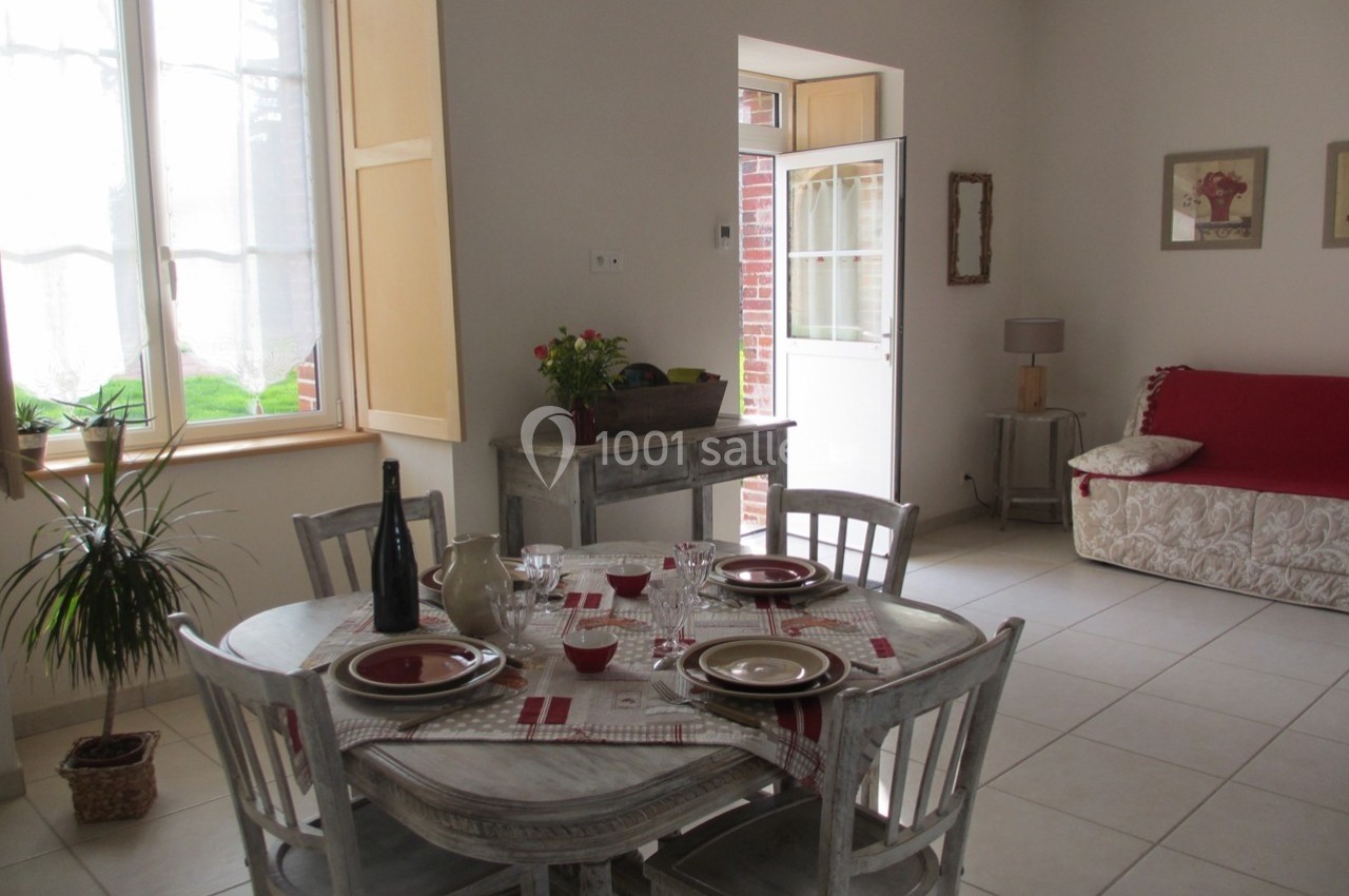 Salle à manger lumineuse avec table dressée, chaises en bois, canapé rouge et porte ouverte sur l'extérieur.