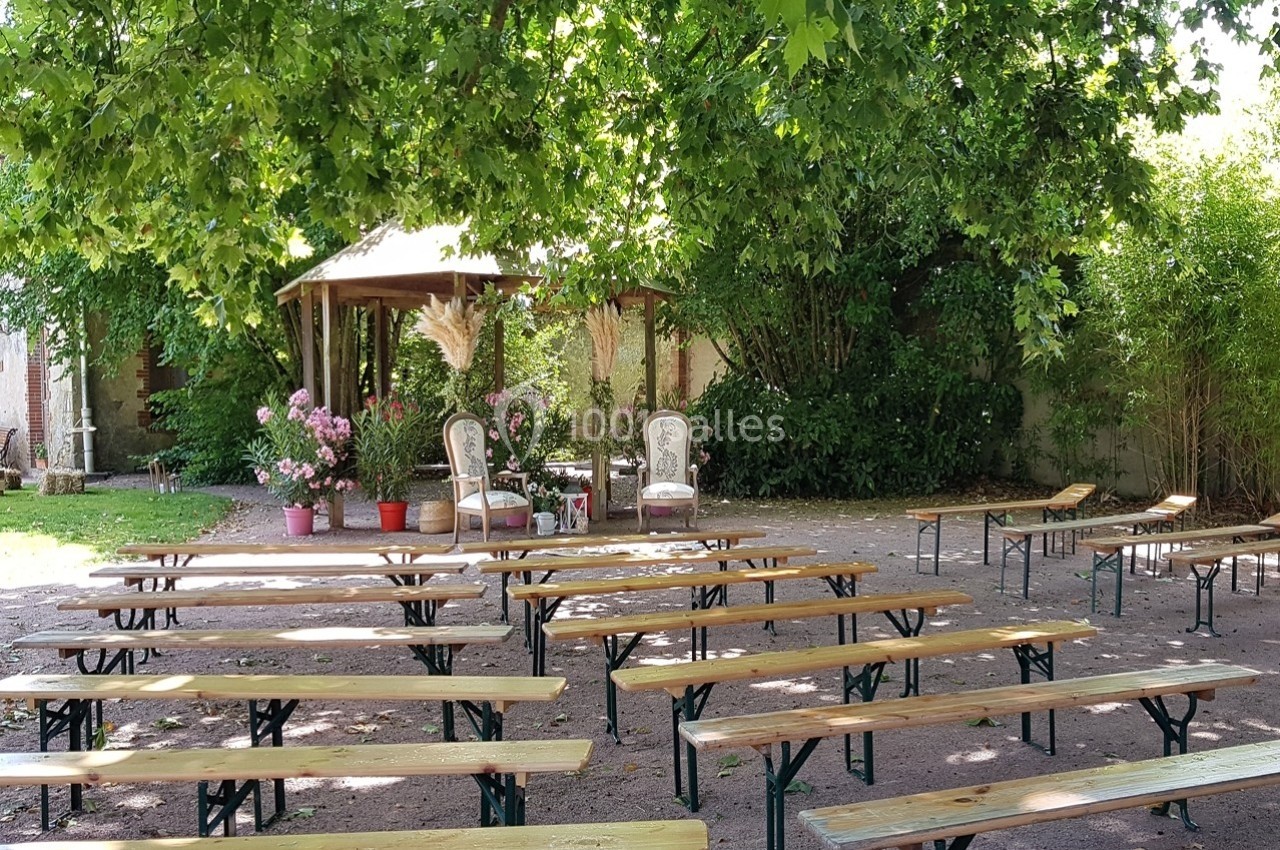 Bancs en bois disposés en extérieur sous des arbres, face à deux fauteuils et une décoration florale.