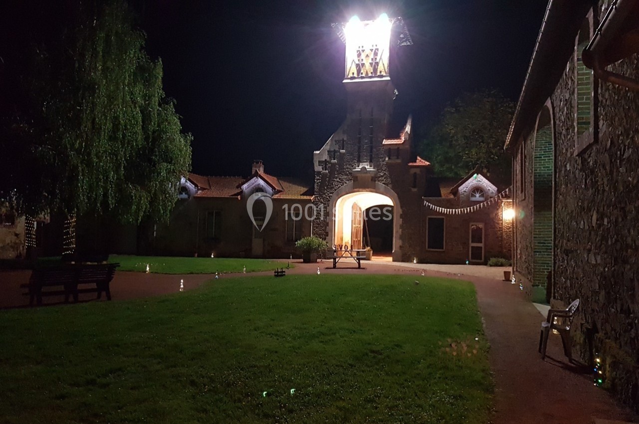Cour éclairée la nuit avec une pelouse, un banc, et un bâtiment en pierre surmonté d'une tour lumineuse.