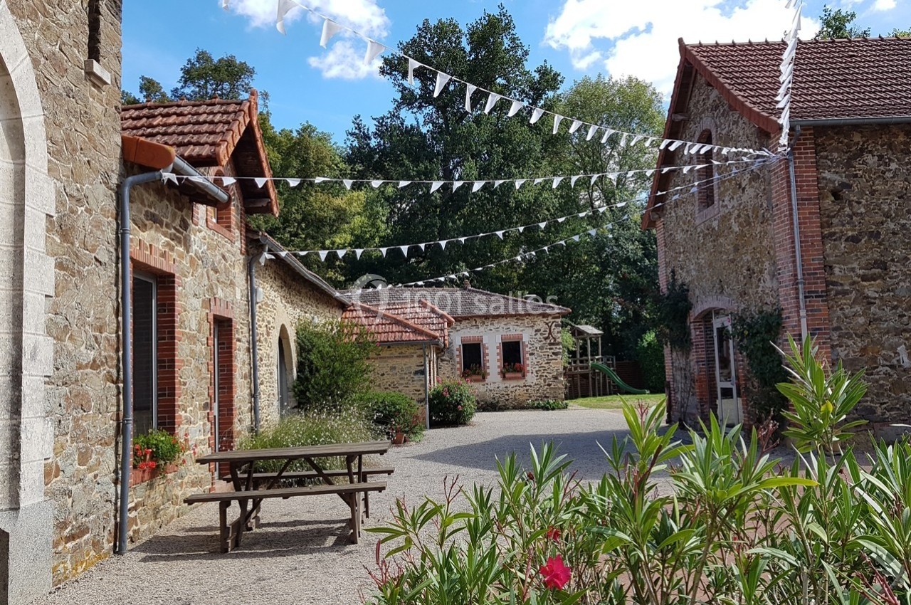 Cour pavée entourée de bâtiments en pierre, décorée de guirlandes blanches, avec une table en bois au premier plan.