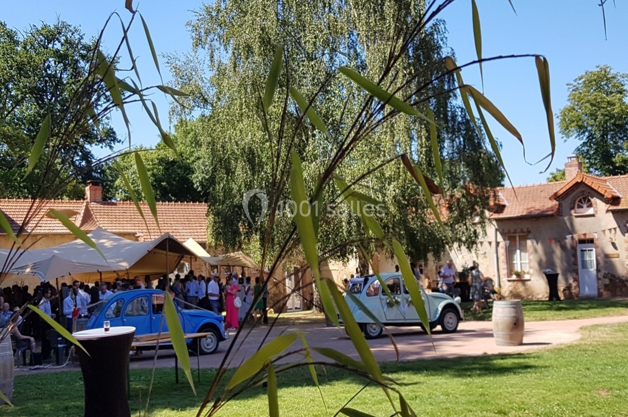 Deux voitures anciennes stationnées devant une maison en pierre, avec des invités rassemblés sous un chapiteau.